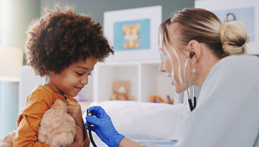 Female doctor using stethoscope on young boy holding teddy bear.